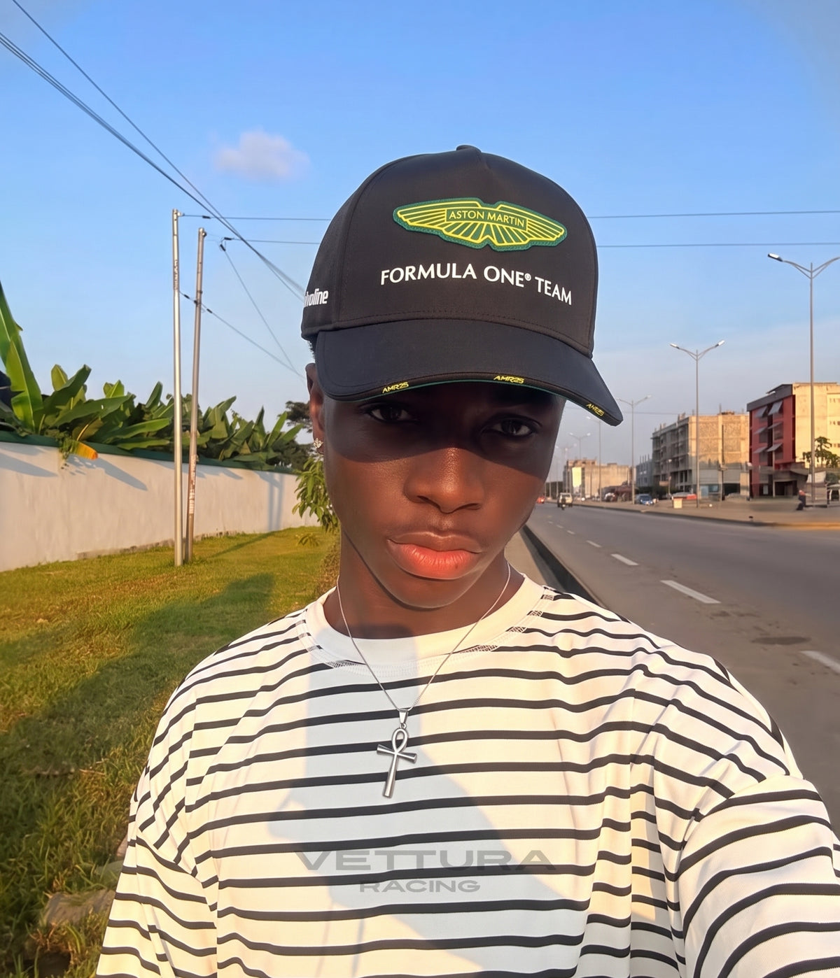 Man in a striped shirt wearing the black Aston Martin F1 Team 2025 cap at sunset