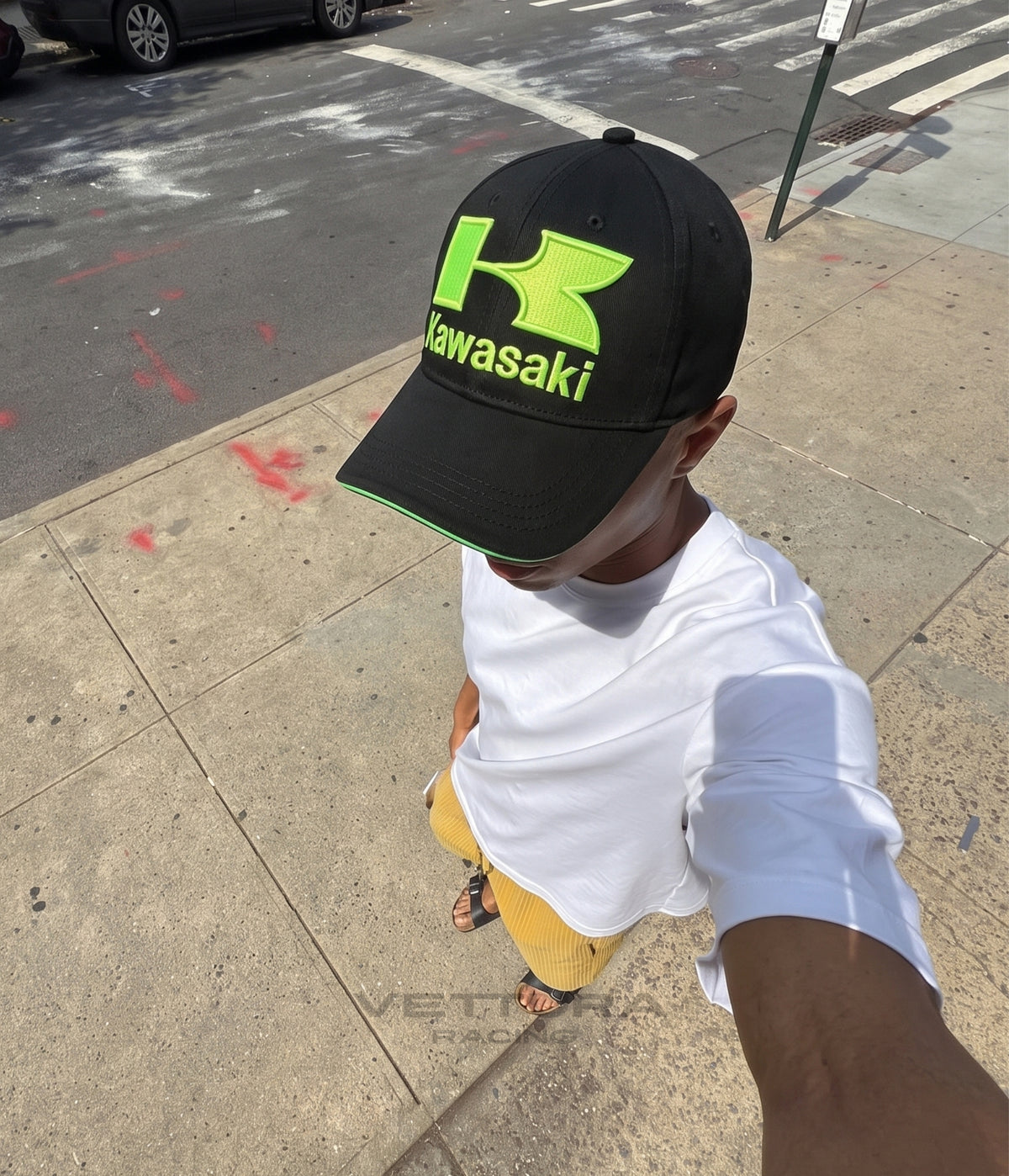 Man in a white tee wearing the black Kawasaki Racing cap on a city sidewalk