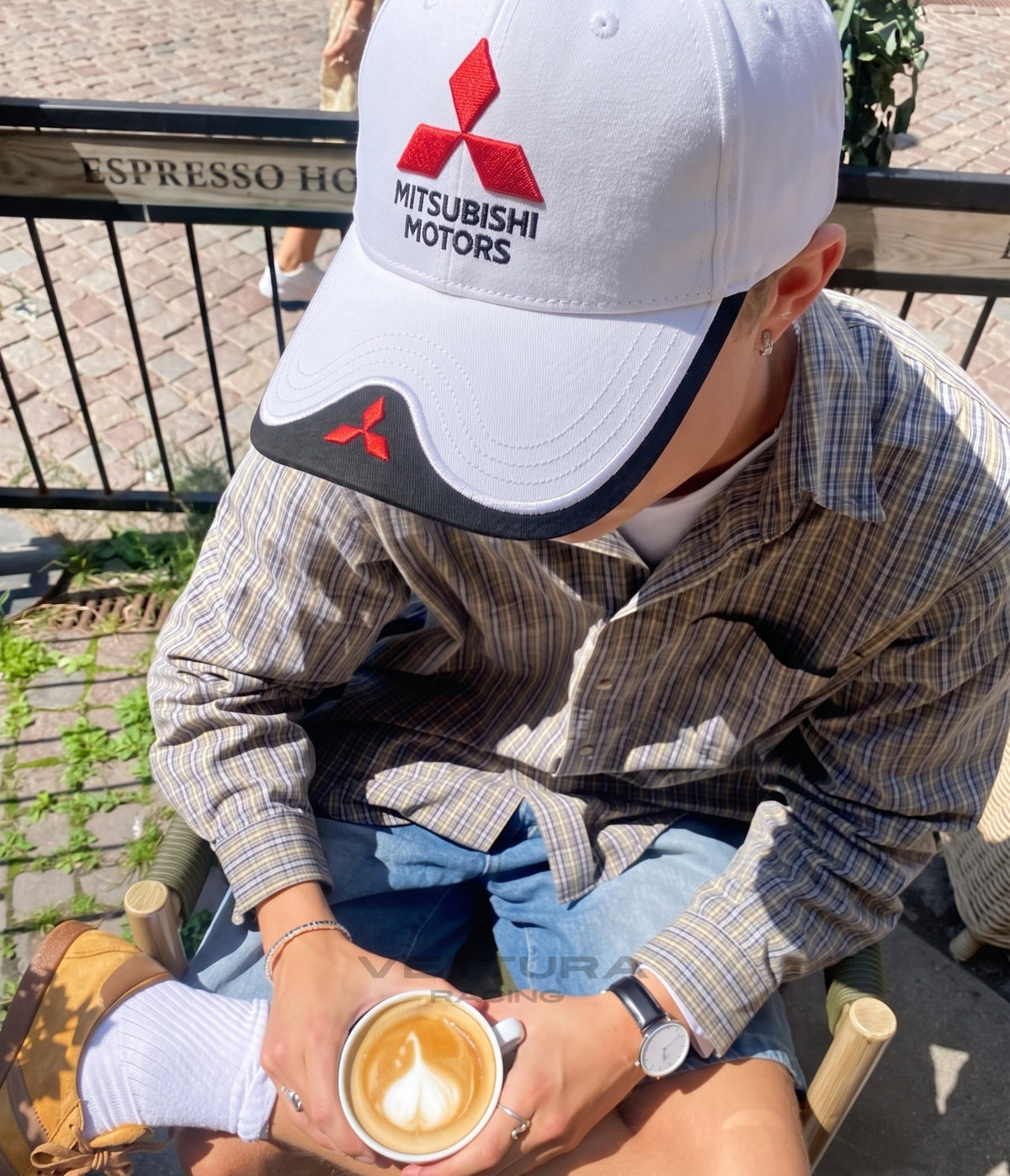 Person in a checked shirt wearing the white Mitsubishi Motors cap at a cafe terrace