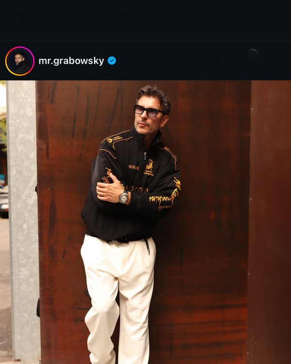 Man in a black vintage racing jacket and cream trousers posing against a rust wall