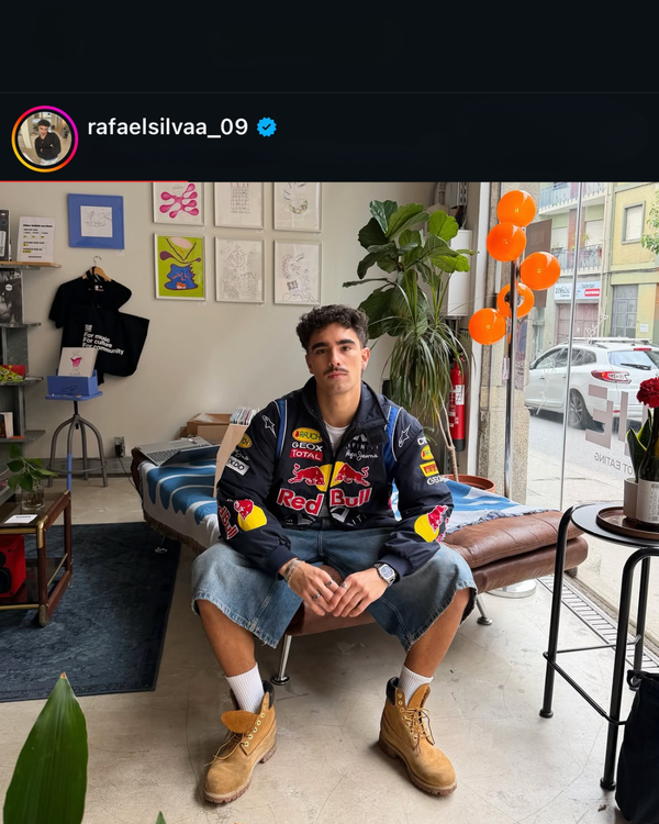 Man seated in a navy Red Bull F1 vintage racing jacket, denim shorts and work boots