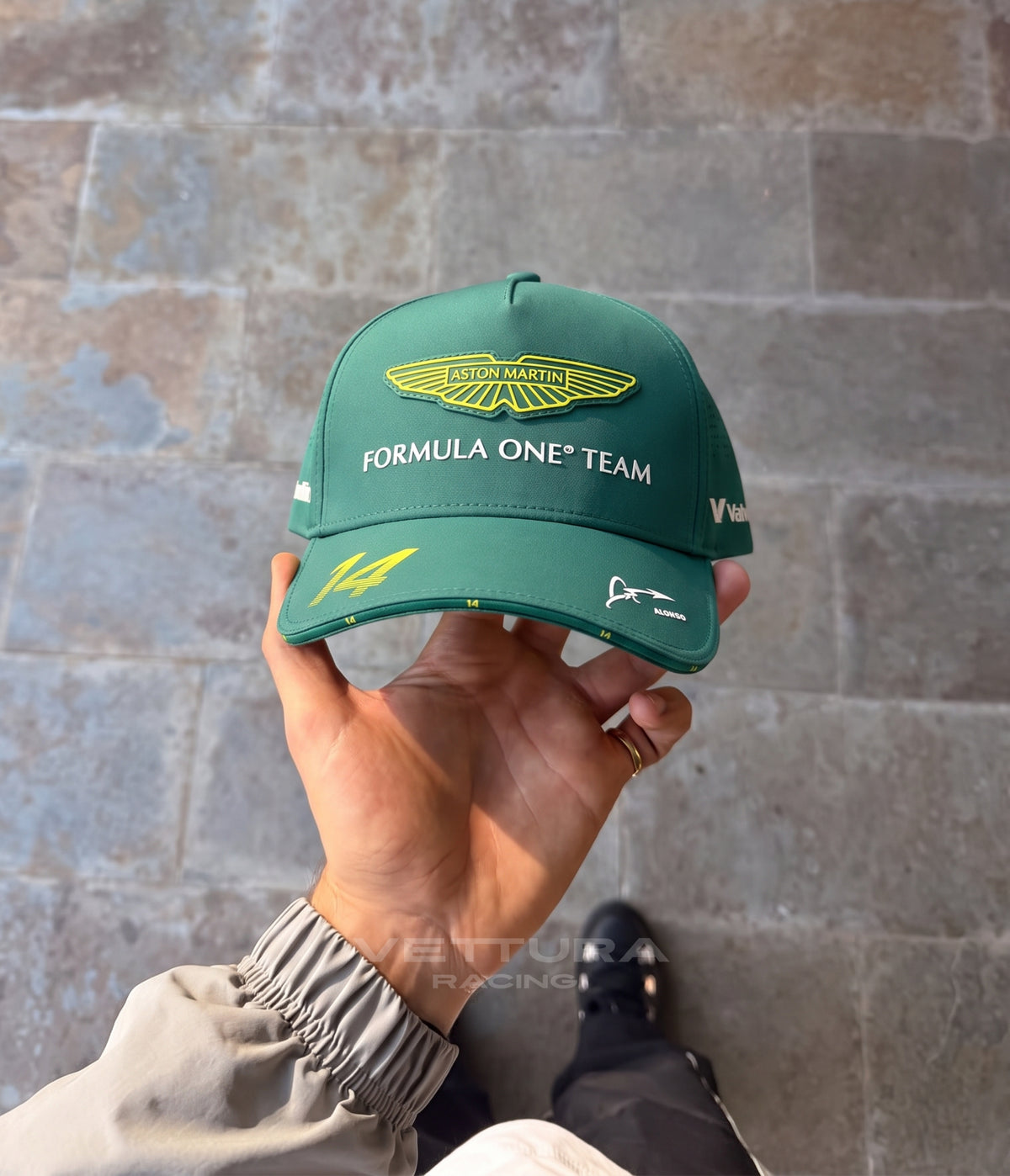 Hand holding the racing green Aston Martin F1 Alonso cap against a stone wall