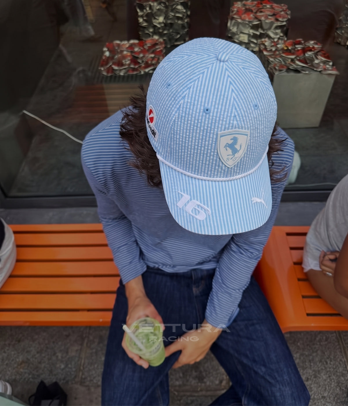 Person wearing the light blue Ferrari F1 seersucker cap seated on an orange bench