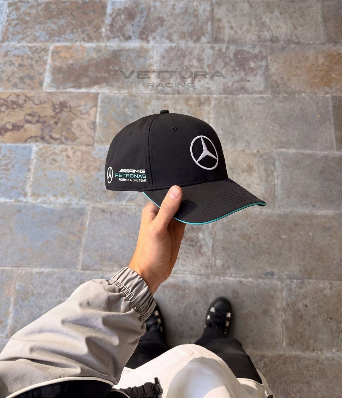 Hand holding the black Mercedes AMG F1 Team cap against a stone wall