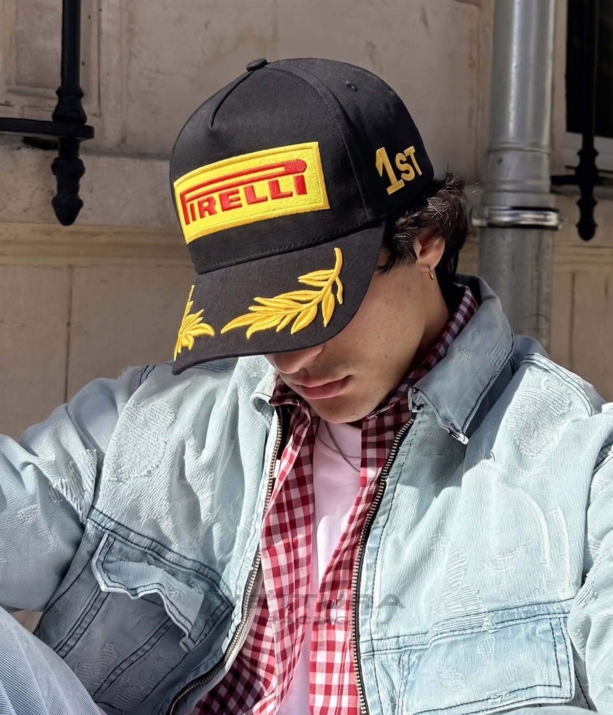 Man in a denim jacket wearing the black Pirelli F1 podium cap with gold laurel brim